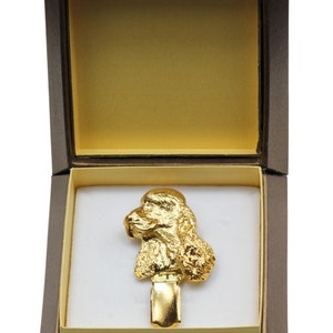 Könnte beinhalten: Eine goldfarbene Pudelfigur in einer braunen Samtbox mit weißem Innenraum. Der Pudel sitzt mit dem Kopf zur Seite geneigt.