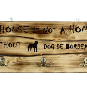 Op de afbeelding: Een houten bord met de tekst "A house is not a home without a Dog de Bordeaux" en drie haken. Het bord is verweerd en heeft een rustieke uitstraling.