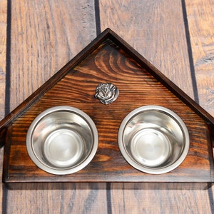Puede incluir: Un soporte de madera para cuencos de comida para mascotas con forma de casa con dos cuencos de acero inoxidable. El soporte es de color marrón oscuro y tiene una rosa de metal decorativa en la parte delantera.