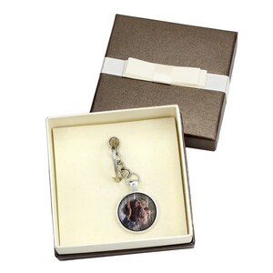 May include: A silver keyring with a round glass charm featuring a brown and white dog. The keyring is in a brown gift box with a white ribbon.