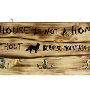 Könnte beinhalten: Ein Holzschild mit dem Text "Ein Haus ist kein Zuhause ohne einen Berner Sennenhund" und einer Silhouette eines Berner Sennenhundes. Das Schild hat drei Haken und ist im rustikalen Stil gealtert.