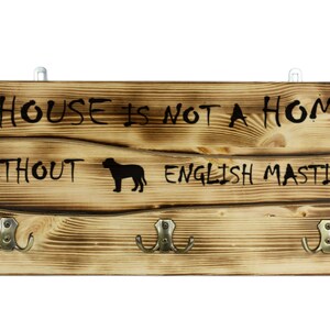 Op de afbeelding: Een houten bord met de tekst "Een huis is geen thuis zonder een Engelse Mastiff" en drie haken. Het bord is donkerbruin gebeitst en heeft een rustieke uitstraling.