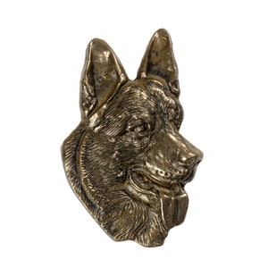 Può includere: Una scultura in metallo dorato della testa di un pastore tedesco. La scultura è dettagliata e realistica, con la lingua del cane che sporge.