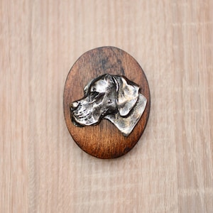 Könnte beinhalten: Ovales Holzschild mit einem metallischen Relief eines Hunde-Kopfes. Das Profil des Hundes ist detailliert, mit Fokus auf Schnauze und Ohr. Das Holz ist dunkelbraun gebeizt.