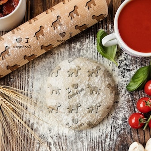 Può includere: Un mattarello di legno con un motivo di cani e il testo "I love Cockapoos" è su una superficie di legno con una palla di pasta, farina, steli di grano, pomodori, basilico, funghi, aglio e una ciotola di salsa di pomodoro.