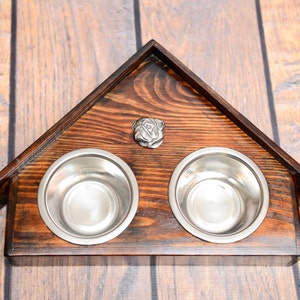 Puede incluir: Un soporte de cuenco para perros de madera con forma de casa con dos cuencos de acero inoxidable y un adorno de metal con forma de cabeza de carlino.