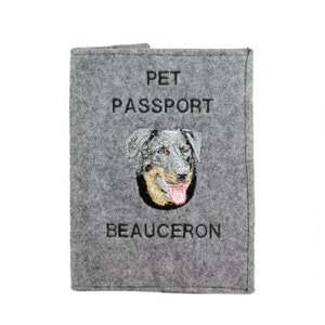 Op de afbeelding: Grijze gevoelde dierenpaspoort houder met de tekst "PET PASSPORT" en "BEAUCERON" geborduurd in zwart. Een geborduurd beeld van een Beauceron hond is in het midden.