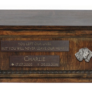 May include: A wooden pet urn with a brown finish. It has a plaque with the inscription "You left our lives, but you will never leave our hearts."  A second plaque reads "Charlie 17.07.2005 † 25.02.2018". A metal dog head is attached to the urn.