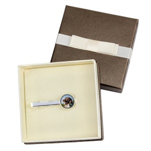 May include: A silver tie clip with a round glass cameo featuring a brown and white dog. The tie clip is in a white and brown gift box with a white satin ribbon.