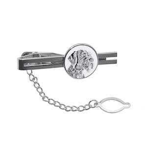 May include: A silver tie clip featuring a black and white image of a dog's head within a round, silver-rimmed frame. The tie clip has a silver chain attached.