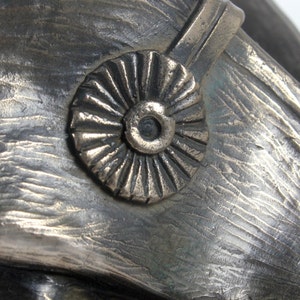 Op de afbeelding: Een close-up van een bronzen sculptuur van een mensenhoofd dat een hoed draagt met een decoratief rond embleem. De hoed heeft een getextureerd oppervlak en een verhoogde band.