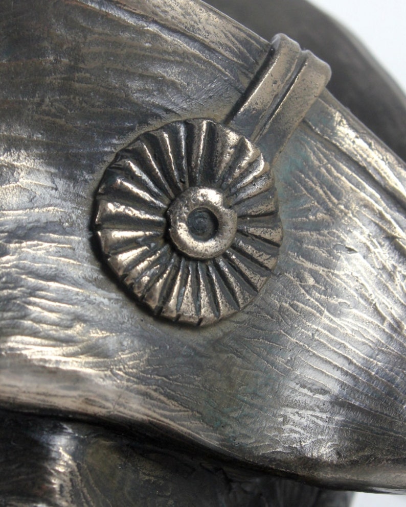 Op de afbeelding: Een close-up van een bronzen sculptuur van een mensenhoofd dat een hoed draagt met een decoratief rond embleem. De hoed heeft een getextureerd oppervlak en een verhoogde band.