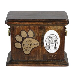 Puede incluir: Una urna de madera para mascotas con un diseño de huella de pata y el texto "You left paw prints on my heart". La urna tiene un retrato de un perro Beagle y una placa de nombre que dice "BUDDY 2005-2018".