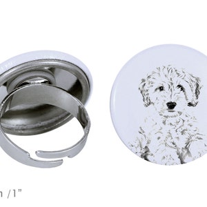 May include: A white ring with a button featuring a black and white illustration of a doodle dog. The ring is 1 inch in diameter.