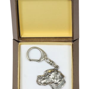 Op de afbeelding: Een zilveren metalen sleutelhanger met een gedetailleerd hondenhoofd, gepresenteerd in een bruine geschenkdoos met een gele binnenkant.