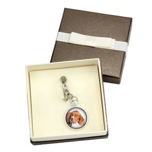 May include: A silver keyring with a circular pendant displaying a dog's portrait, presented in a brown gift box with a cream ribbon. The dog has brown and white fur.