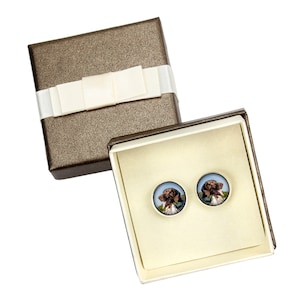 May include: A pair of brown and white dog portrait earrings in a cream-colored gift box with a brown lid and a white satin bow.