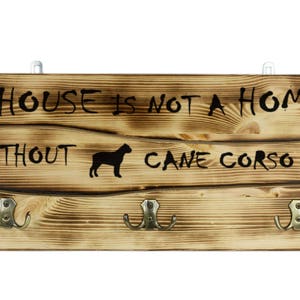 Op de afbeelding: Houten wandkapstok met drie haken. De kapstok heeft de tekst "A HOUSE IS NOT A HOME WITHOUT CANE CORSO" in zwarte letters en een silhouet van een hond. Het hout heeft een natuurlijke, rustieke afwerking.