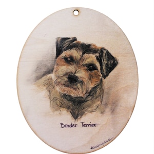 May include: A hand-painted wooden ornament featuring a Border Terrier dog. The dog is depicted in a realistic style with brown and white fur. The ornament is oval-shaped and has a hole at the top for hanging. The text "Border Terrier" is written below the dog's image.