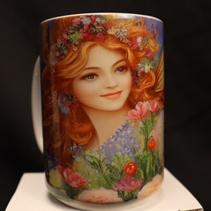 May include: White ceramic mug featuring a colourful image of a woman with long red hair wearing a flower crown. The woman is surrounded by flowers and greenery.