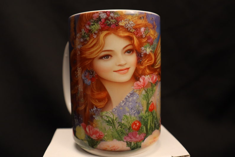 May include: White ceramic mug featuring a colourful image of a woman with long red hair wearing a flower crown. The woman is surrounded by flowers and greenery.