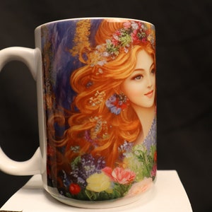 May include: White ceramic mug featuring a colourful image of a woman with long red hair wearing a flower crown. The image is surrounded by flowers and greenery.