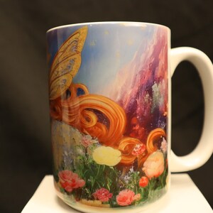 May include: White ceramic mug featuring a colourful image of a fairy with golden wings and long red hair. The fairy is flying over a field of flowers with a mountain range in the background.