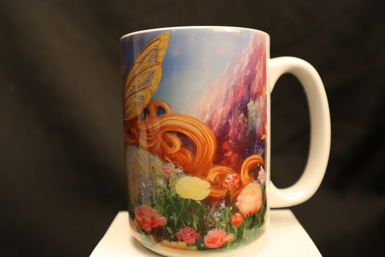 May include: White ceramic mug featuring a colourful image of a fairy with golden wings and long red hair. The fairy is flying over a field of flowers with a mountain range in the background.