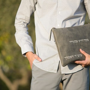 May include: A gray leather clutch with a zipper closure and silver Hebrew text. The text reads "ברוך הבא הביתה" and "שבת שלום".
