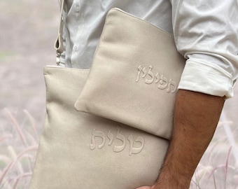 Puede incluir: Dos bolsas de cuero blanquecino, una más grande que la otra, con texto hebreo en relieve. Las bolsas son sostenidas por una persona que lleva una camisa blanca. El fondo es una vista borrosa de hierba alta.