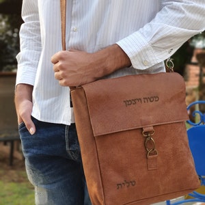 May include: A brown leather messenger bag with a flap closure and a metal clasp. The bag has a personalized inscription in Hebrew that reads "Sarah Yisakov" and "Shlomit".
