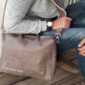 May include: A brown leather messenger bag with a strap and a white inscription that reads "מסע מצליח תמיד".