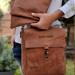 May include: A brown leather messenger bag with a flap closure and a metal clasp. The bag has three smaller brown leather pouches with Hebrew text on them. The bag is worn over the shoulder of a person wearing a white striped shirt and blue jeans.