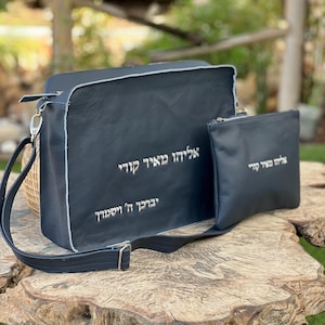 May include: A black leather messenger bag and matching pouch with white stitching and silver text. The bag has a shoulder strap and is sitting on a wooden surface. The text on the bag is in Hebrew.