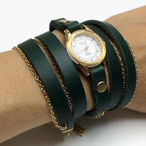 May include: A gold-tone watch with a white face and black hands is wrapped in a dark green leather band. The band is wrapped around the wrist multiple times and has a gold-tone buckle.