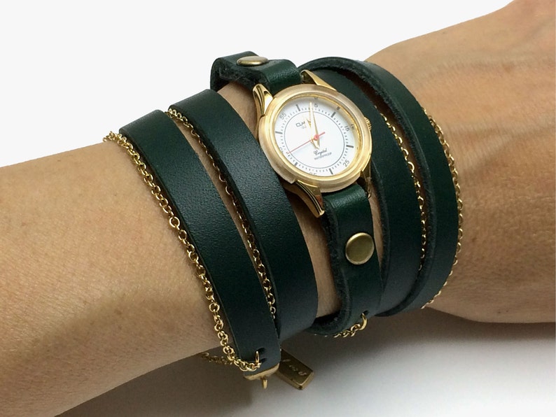 May include: A gold-tone watch with a white face and black hands is wrapped in a dark green leather band. The band is wrapped around the wrist multiple times and has a gold-tone buckle.
