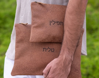 Puede incluir: Dos bolsas de cuero marrón con cremalleras. La bolsa más grande tiene la palabra "שלום" (hebreo para "paz") escrita en ella. La bolsa más pequeña tiene la palabra "תפילה" (hebreo para "oración") escrita en ella.