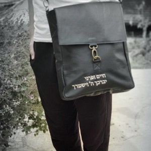 May include: A black leather messenger bag with a gold clasp and a Hebrew inscription that reads "Here I am, I am here".