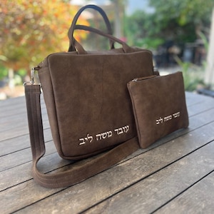 May include: A brown suede-like shoulder bag with a matching small pouch. The bag has a top handle and a detachable shoulder strap. The text "עובד משה ליב" is embroidered on both the bag and the pouch.