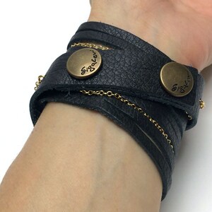 Op de afbeelding: Zwarte leren armband met een gouden ketting en twee knoopsluitingen van messing. De knopen zijn gegraveerd met de tekst "Siglo Levi".