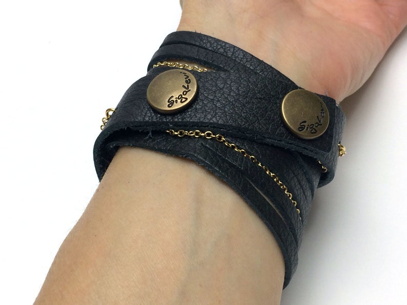 Op de afbeelding: Zwarte leren armband met een gouden ketting en twee knoopsluitingen van messing. De knopen zijn gegraveerd met de tekst "Siglo Levi".