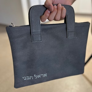 May include: A gray leather bag with a zipper closure and two top handles. The bag has the Hebrew words "אֲדַרְתָּ"  "תַּחְבִּיבִּי" embroidered on the front.