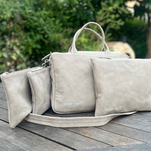 May include: Set of four beige suede bags with zipper closures. The largest bag has a top handle and a shoulder strap. Each bag has Hebrew text embroidered on the front.