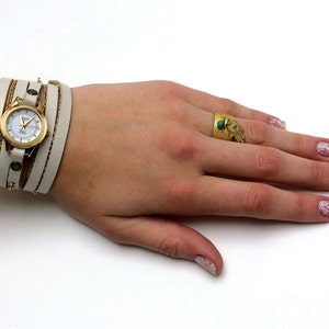 May include: A white leather wrap watch with a gold face and gold chain accents. The watch is on a person's wrist. The person is also wearing a gold ring with a turquoise stone on their finger.