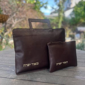 May include: Two brown leather bags with gold Hebrew lettering. The larger bag has a handle and the smaller bag has a zipper. The lettering on the larger bag reads "Ma'avod Yachid" and the lettering on the smaller bag reads "Ma'avod Yachid".