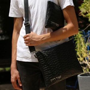 May include: A black leather crossbody bag with a crocodile embossed pattern. The bag has a silver zipper and a silver strap. The bag has the Hebrew text "כבוד יהוה" written on it.