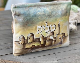 Puede incluir: Un bolso de mano rectangular de cuero blanquecino con cierre de cremallera. El bolso presenta una escena pintada a mano de una ciudad con cúpulas y una torre, sobre un cielo amarillo y azul. La palabra "Jerusalén" está escrita en hebreo.