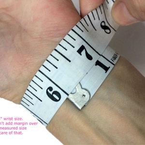Op de afbeelding: Een witte meetlint gewikkeld om een pols, met een meting van 6 inch. De tekst "This is a 6" wrist size. Please don't add margin over the tight measured size as I take care of that." staat onder de afbeelding.