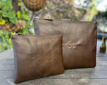 Puede incluir: Dos bolsas de cuero marrón con cierres de cremallera. La bolsa más grande tiene texto hebreo bordado. La bolsa más pequeña también tiene texto hebreo bordado. Las bolsas están sobre una superficie de madera.