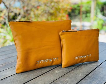 Puede incluir: Dos bolsas de cuero color mostaza con texto plateado. La bolsa más grande es rectangular y la más pequeña cuadrada. Ambas tienen cierre de cremallera. Las bolsas están sobre una superficie de madera.